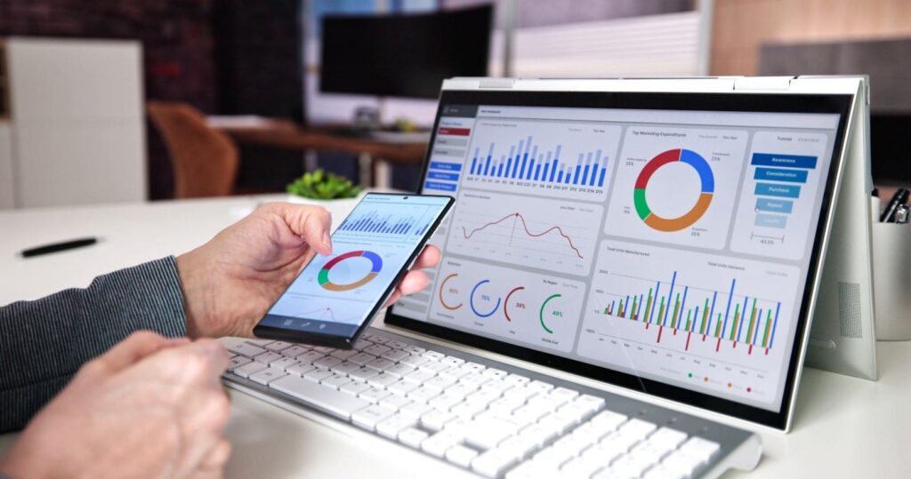 Primo piano di un pc con visuale di dashboard di un gestionale; si vede una mano che tiene uno smartphone con lo stesso programma sullo schermo