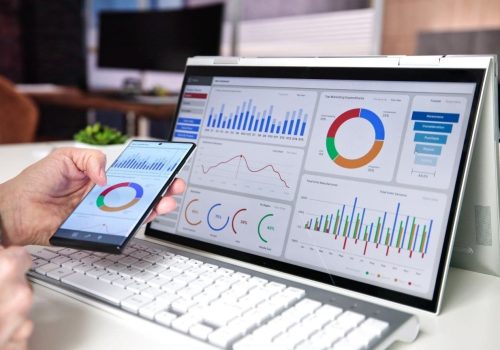 Primo piano di un pc con visuale di dashboard di un gestionale; si vede una mano che tiene uno smartphone con lo stesso programma sullo schermo